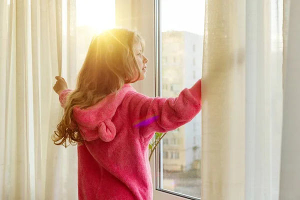Bonjour. Bonjour. Fille enfant en pyjama ouvre les rideaux et regarde par la fenêtre — Photo