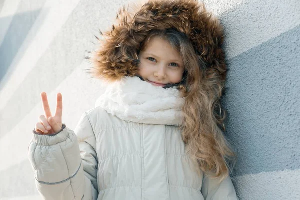 Κοντινό στο υπαίθριο χειμερινό πορτρέτο του παιδιού, ξανθιά κορίτσι με σγουρά μαλλιά 7, 8 χρόνια σε γούνα κουκούλα, κορίτσι χαμογελάει δείχνοντας σημάδι νίκης — Φωτογραφία Αρχείου
