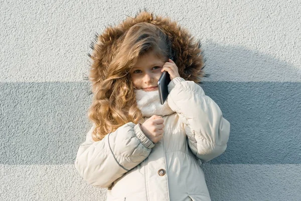 Primer plano al aire libre retrato de invierno de niño, chica rubia con el pelo rizado de 7, 8 años en la capucha de piel, chica sonriendo mostrando signo de victoria, sonriendo hablando por teléfono móvil — Foto de Stock