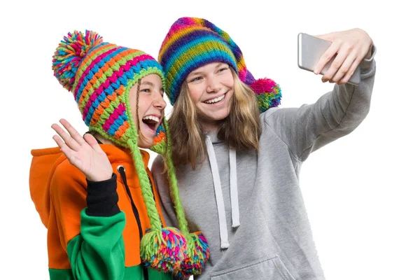 Vinter porträtt av två lyckliga vackra tonårs flickvänner i stickade hattar ha kul med mobiltelefon, ta selfie, isolerad på vit bakgrund. Människor, ungdom och vänskap koncept — Stockfoto