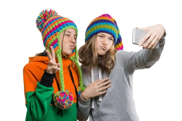 Vinter porträtt av två lyckliga vackra tonårs flickvänner i stickade hattar ha kul med mobiltelefon, ta selfie, isolerad på vit bakgrund. Människor, ungdom och vänskap koncept — Stockfoto