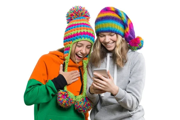Retrato de inverno de duas belas amigas adolescentes felizes em chapéus de malha se divertindo com telefone celular, isolado no fundo branco. Pessoas, juventude e conceito de amizade — Fotografia de Stock