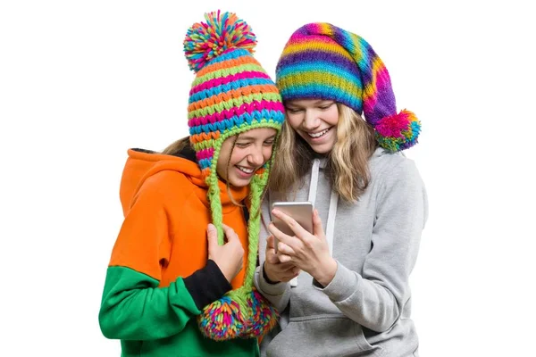 Vinter porträtt av två glada leende vackra flickor i stickade hattar ha kul med mobiltelefon, isolerad på vit bakgrund. Människor, ungdom och vänskap koncept — Stockfoto