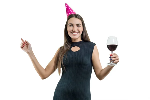 Felice giovane donna sorridente con un bicchiere di vino rosso, in cappelli da festa, abito da cocktail nero. Su sfondo bianco isolato ragazza divertirsi ballando — Foto Stock