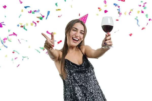 Chica morena elegante y sonriente en sombrero de fiesta con copa de vino tinto bailando bajo confeti y riéndose, fondo blanco aislado. Fiesta, bebidas, vacaciones y concepto de celebración . —  Fotos de Stock