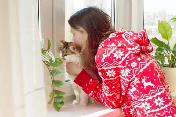 Wintertijd thuis, jong meisje in de winter warme pyjama met een kat die uitkijkt op het raam — Stockfoto