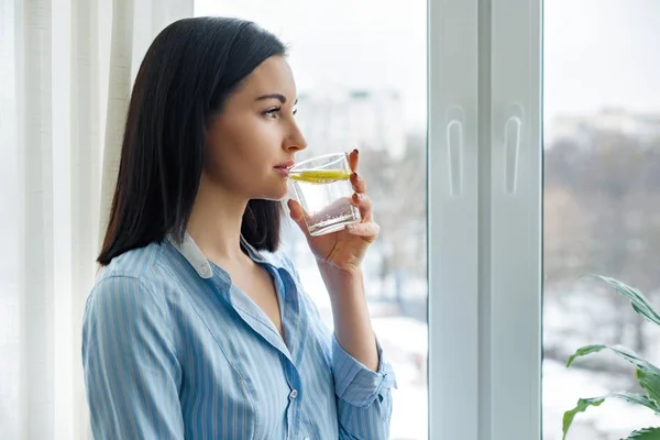 Jeune femme matin à la maison près de la fenêtre boire de l'eau avec du citron, boisson vitaminée en hiver printemps saison — Photo