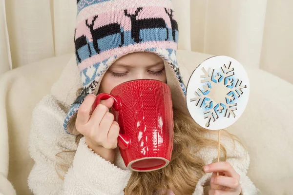 Klein meisje zittend thuis in de stoel, in gebreide muts met kerst ornament met kopje warme drank. Meisje vasthouden symbolische Snowflake — Stockfoto