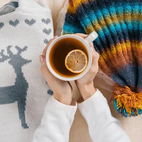 Winter is coming, womens hands are holding cup of hot tea with lemon, background is warm seasonal clothes, close-up view from above — Stock Photo, Image