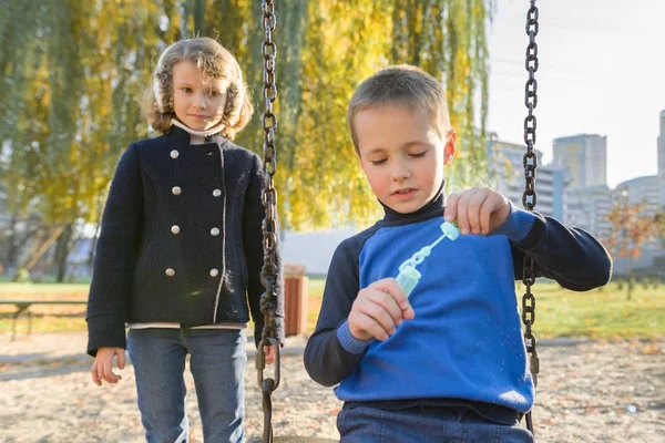 Маленький мальчик и девочка играют в осеннем парке, дети сидят на качелях мыльные пузыри — стоковое фото