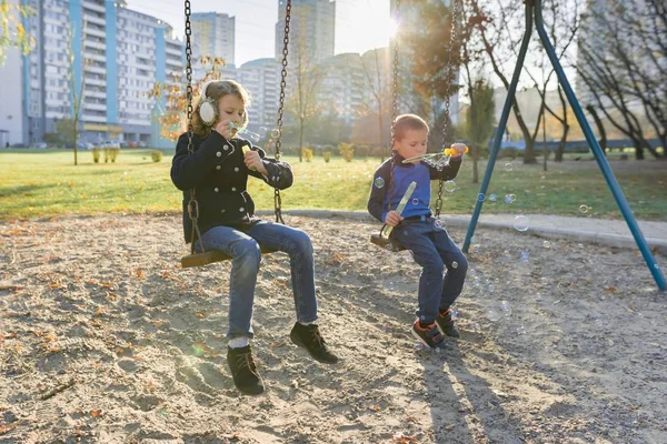 Little boy and girl playing in autumn park, children sitting on swing blow soap bubbles — Stock Photo, Image