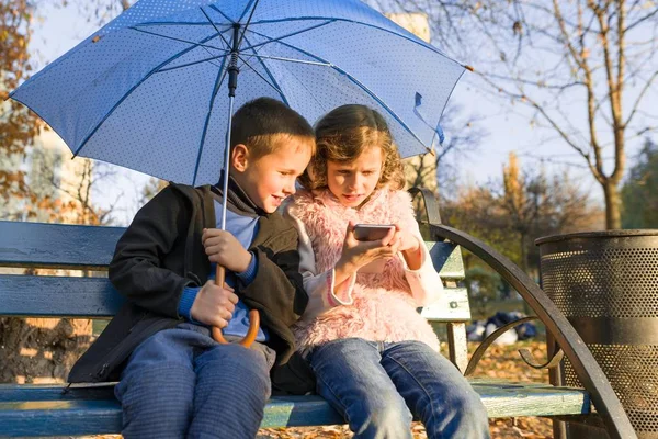 Дети сидят на скамейке в осеннем парке, используя смартфон — стоковое фото