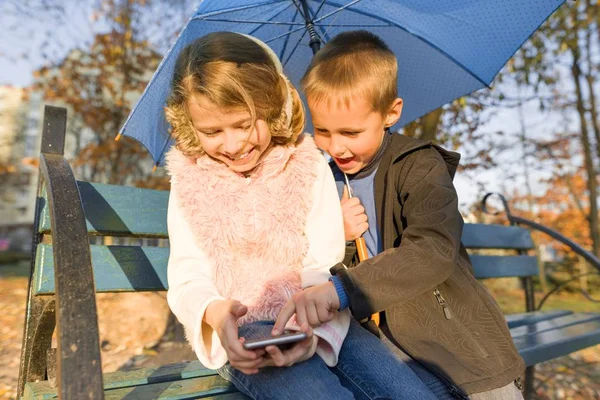 Dzieci siedzą na ławce w jesiennym parku, za pomocą smartfona — Zdjęcie stockowe