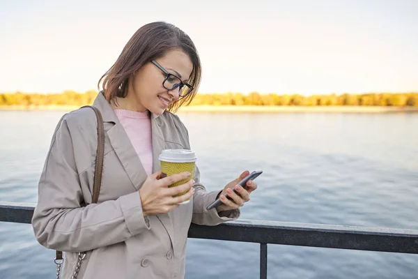 Retrato de mulher ao ar livre dos anos 40, feminino sorridente com xícara de café e smartphone — Fotografia de Stock