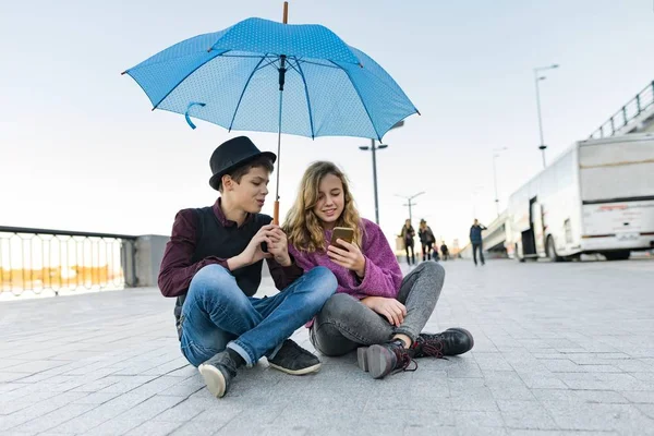 Freunde des Paares sitzen unter einem Regenschirm und schauen aufs Smartphone — Stockfoto