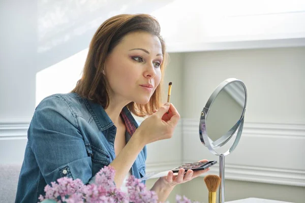 Mogen kvinna tittar på hennes ansikte i spegeln, gör makeup — Stockfoto