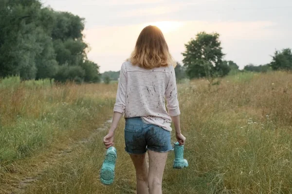 Chica adolescente caminando por el camino rural con botas en las manos —  Fotos de Stock