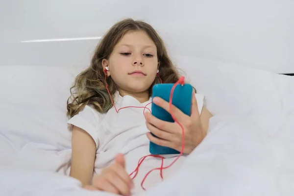 Kislány otthon ül az ágyban, okostelefonnal és fülhallgatóval — Stock Fotó