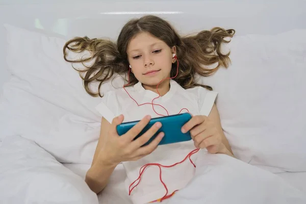 Niña sentada en casa en la cama, con smartphone y auriculares — Foto de Stock