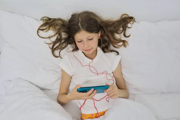 Девочка ребенок с мобильным телефоном в наушниках, сидя в доме белая кровать — стоковое фото