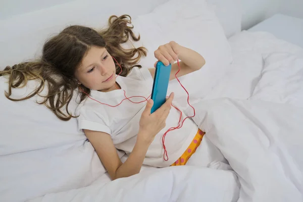 Niña con teléfono móvil en auriculares, sentada en casa cama blanca — Foto de Stock