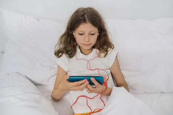 Маленькая девочка сидит дома в постели, со смартфоном и наушниками — стоковое фото