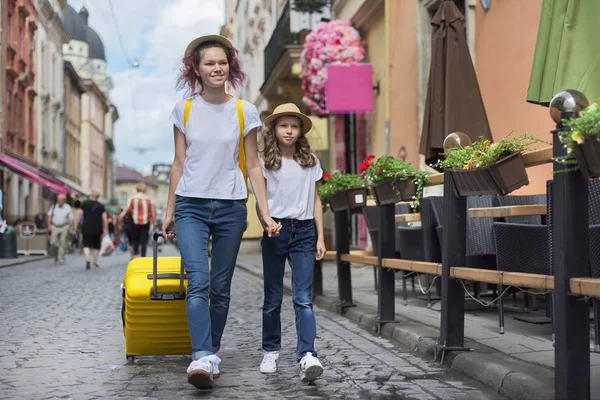 Dos chicas caminando por la ciudad turística cogidas de la mano con una maleta amarilla — Foto de Stock