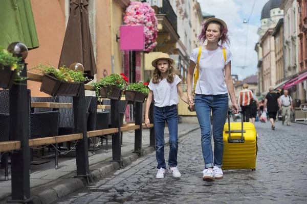 Dos chicas caminando por la ciudad turística cogidas de la mano con una maleta amarilla — Foto de Stock