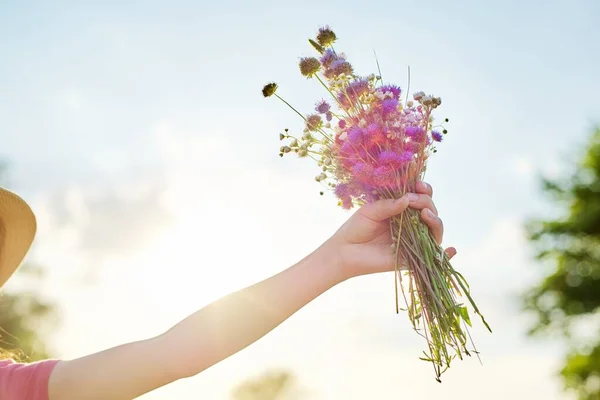 Bouquet of summer wildflowers in girls hand, sky in clouds background, sunset sun lighting — Stock Photo, Image