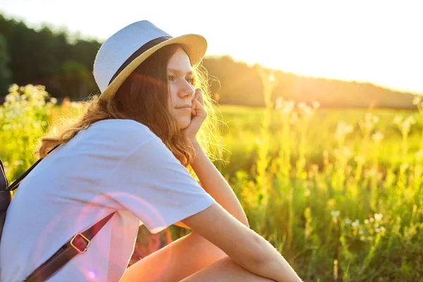 Feliz chica adolescente en sombrero con mochila sentada en el camino del campo, disfrutando de las vacaciones de verano —  Fotos de Stock