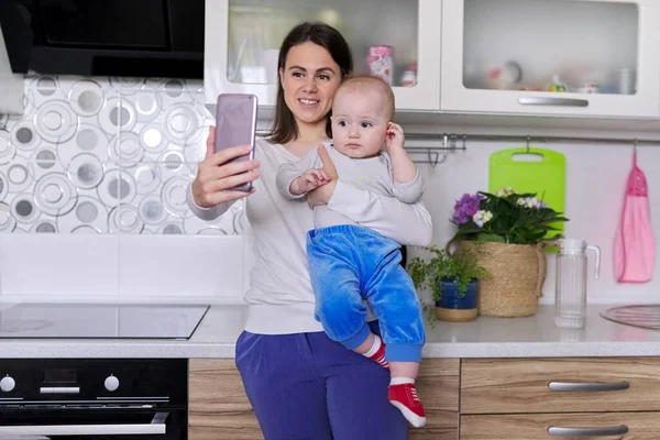 Молодая улыбающаяся женщина с малышом в руках делает видеозвонок с помощью смартфона — стоковое фото
