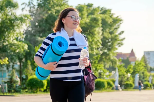 Utomhus glad mogen kvinna promenader efter passform övningar i park, med ryggsäck, yogamatta — Stockfoto