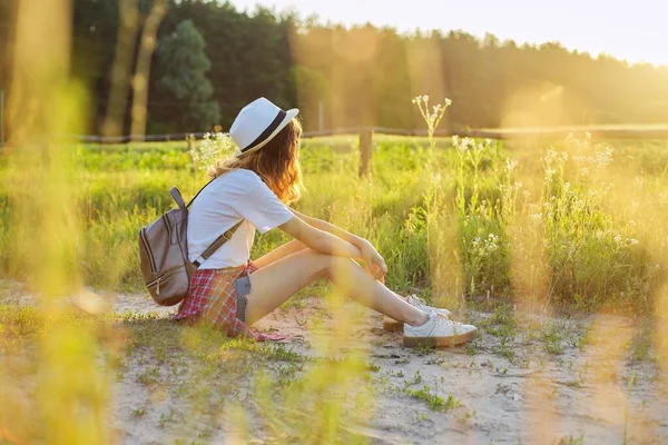 Felice adolescente in cappello con lo zaino seduto sulla strada di campagna, godendo le vacanze estive — Foto Stock