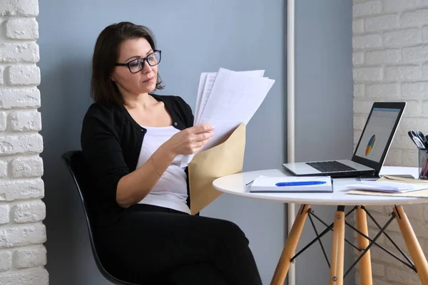 Dojrzała bizneswoman z papierową kopertą siedzi przy biurku z laptopem — Zdjęcie stockowe