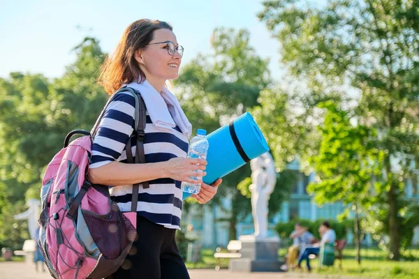 На відкритому повітрі щаслива зріла жінка ходить після вправ у парку, з рюкзаком, йога килимок — стокове фото