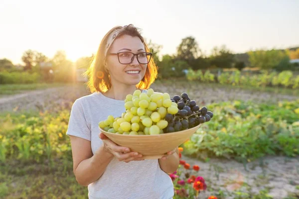 Vrouw met schaal van vers geplukte blauwe en groene druiven, zonnige tuin achtergrond — Stockfoto