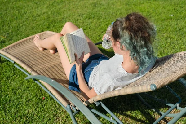 Дівчина-підліток розслабляється на відкритому повітрі, читає книгу і п'є воду — стокове фото