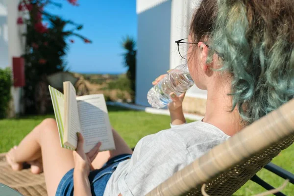 Teenager dívka odpočívající v venkovní židli na trávníku, čtení knihy, pije vodu z láhve — Stock fotografie