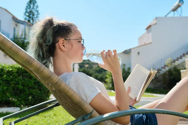 Девочка-подросток отдыхает в открытом кресле на лужайке, читает книги, пьет воду из бутылки — стоковое фото