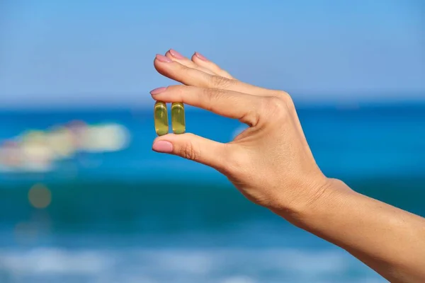 Vitamin D, capsule with fish oil in hand close-up, blue sky sea background