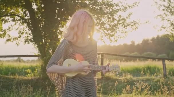 Adolescente chica juega en ukelele, hipster con el pelo teñido de color descansando sentado en la hierba con la guitarra hawaii — Vídeo de stock