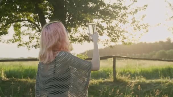 Adolescente chica sosteniendo vidrio con agua mineral pura transparente, puesta de sol sol naturaleza fondo — Vídeo de stock