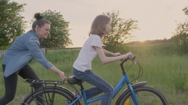 Mamá enseña a la hija del niño a montar en bicicleta, niña monta bicicleta en el camino del campo, puesta del sol paisaje natural fondo . — Vídeos de Stock