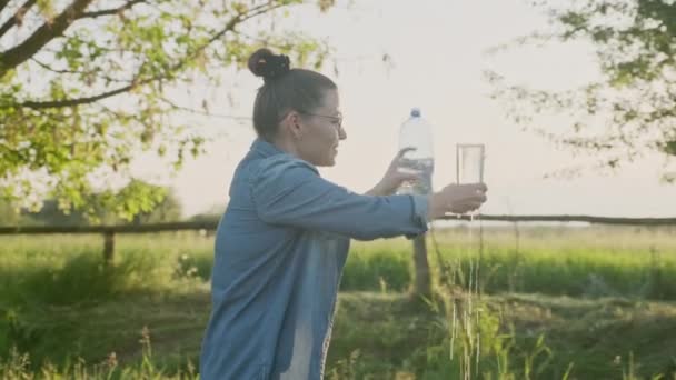 Frau gießt Mineralwasser aus Flasche in Glas, Frau trinkt mit Vergnügen klares Wasser, Sonnenuntergang Sonne Natur Hintergrund — Stockvideo