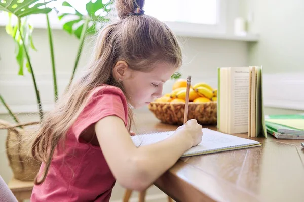 Niña estudiando en casa, sentada en el escritorio, escribiendo en el cuaderno de la escuela — Foto de Stock