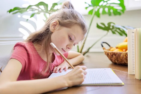 Niña estudiando en casa, sentada en el escritorio, escribiendo en el cuaderno de la escuela — Foto de Stock