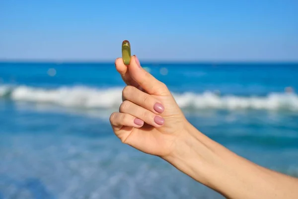 Vitamin D, capsule with fish oil in hand close-up, blue sky sea background — Stock Photo, Image