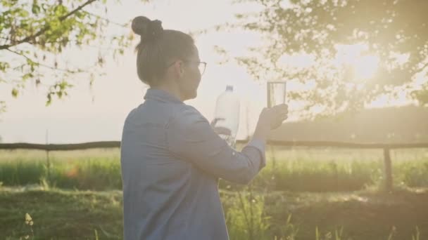 Frau gießt Mineralwasser aus Flasche in Glas, Frau trinkt mit Vergnügen klares Wasser, Sonnenuntergang Sonne Natur Hintergrund — Stockvideo