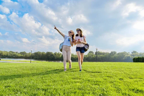 Due ragazze adolescenti che camminano insieme sul prato con bevande nella soleggiata giornata estiva — Foto Stock