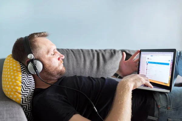 Человек с ноутбуком дома, мужчина расслабляется лежа на диване глядя на монитор ноутбука — стоковое фото